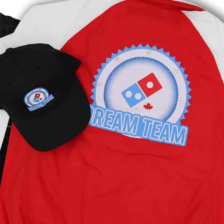 Embroidery Services A demo of corporate apparel featuring a jacket and a hat. The items, a black jacket and a red baseball cap, both have the Domino's Pizza logo custom embroidered on them. The detailed stitching on both the jacket and hat showcases the quality and precision of the corporate customization.